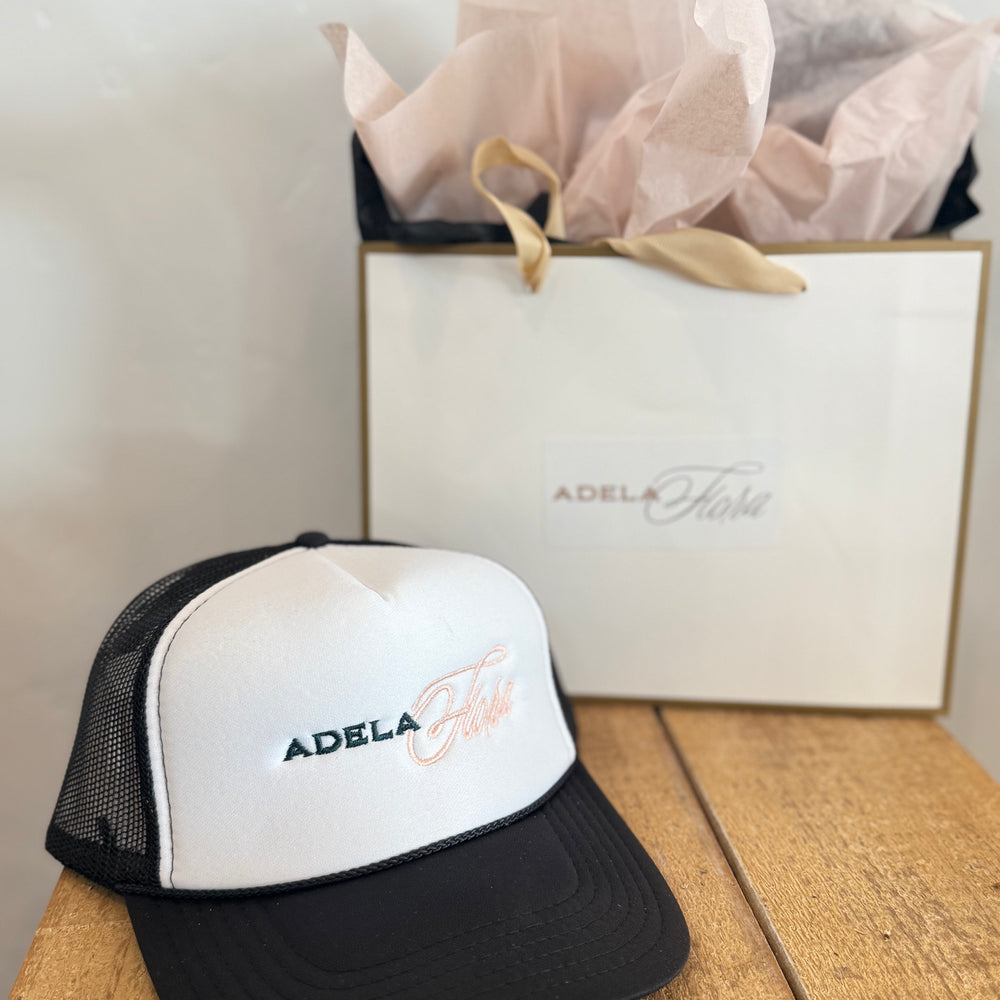 Black and white cap with 'Adela' branding on a wooden surface next to a branded box.
