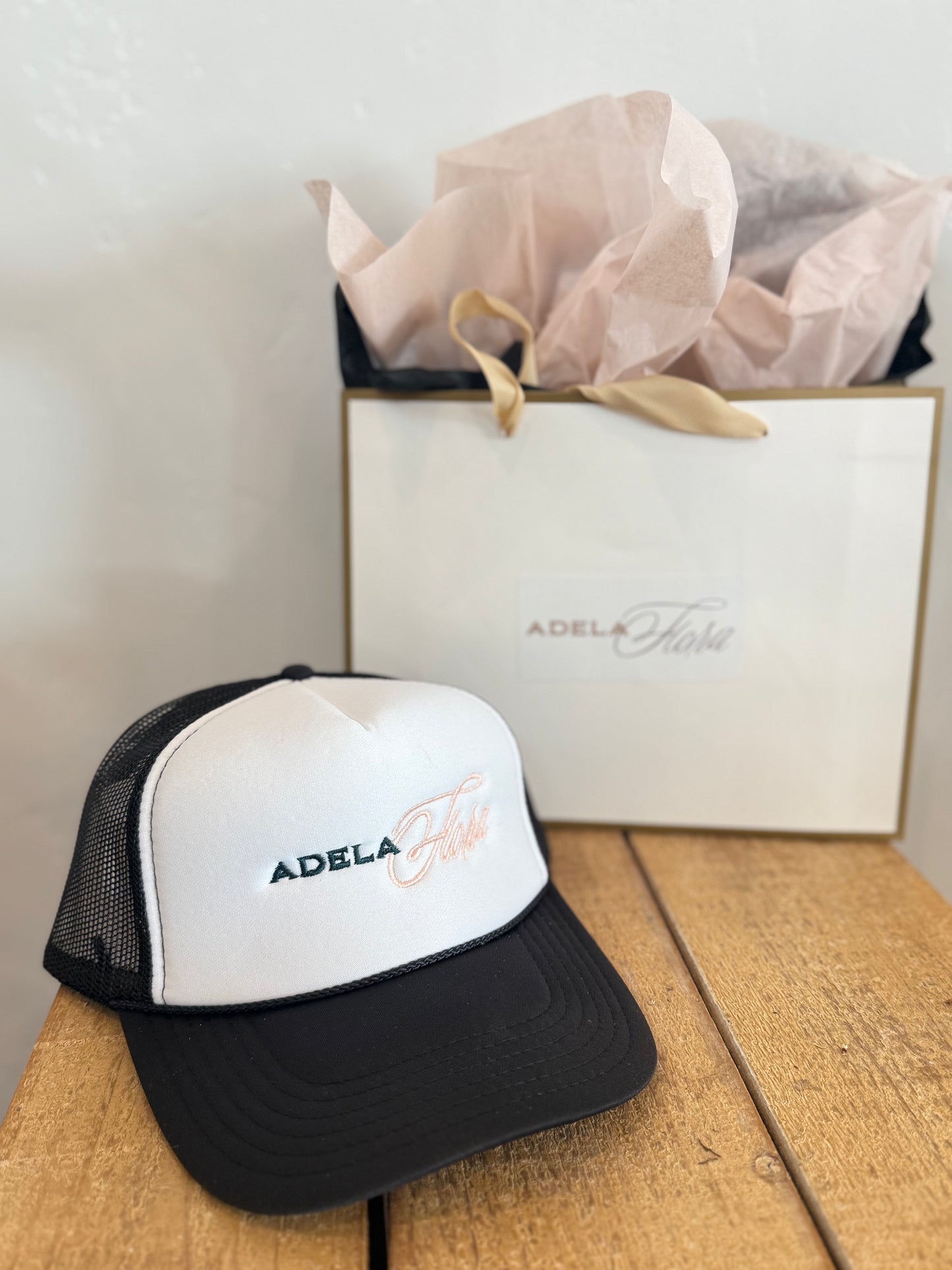 Black and white cap with 'Adela' branding on a wooden surface next to a branded box.